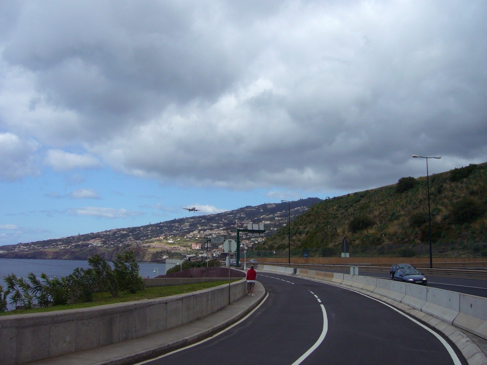 An airliner on final approach to Madeira Airport under heavy cloud, seen from the coastal expressway with the terraced houses of Santa Cruz rising behind.
