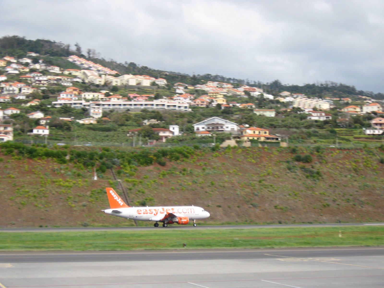 An Airbus A319 touching down at Madeira Airport, with the terraced hillside and pastel houses of Santa Cruz rising steeply behind the runway embankment.