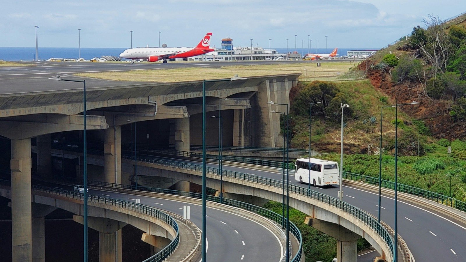 Madeira Airport from above the road interchange, two airliners on the apron beside the control tower, the runway carried on concrete pillars in the foreground and the Atlantic behind.