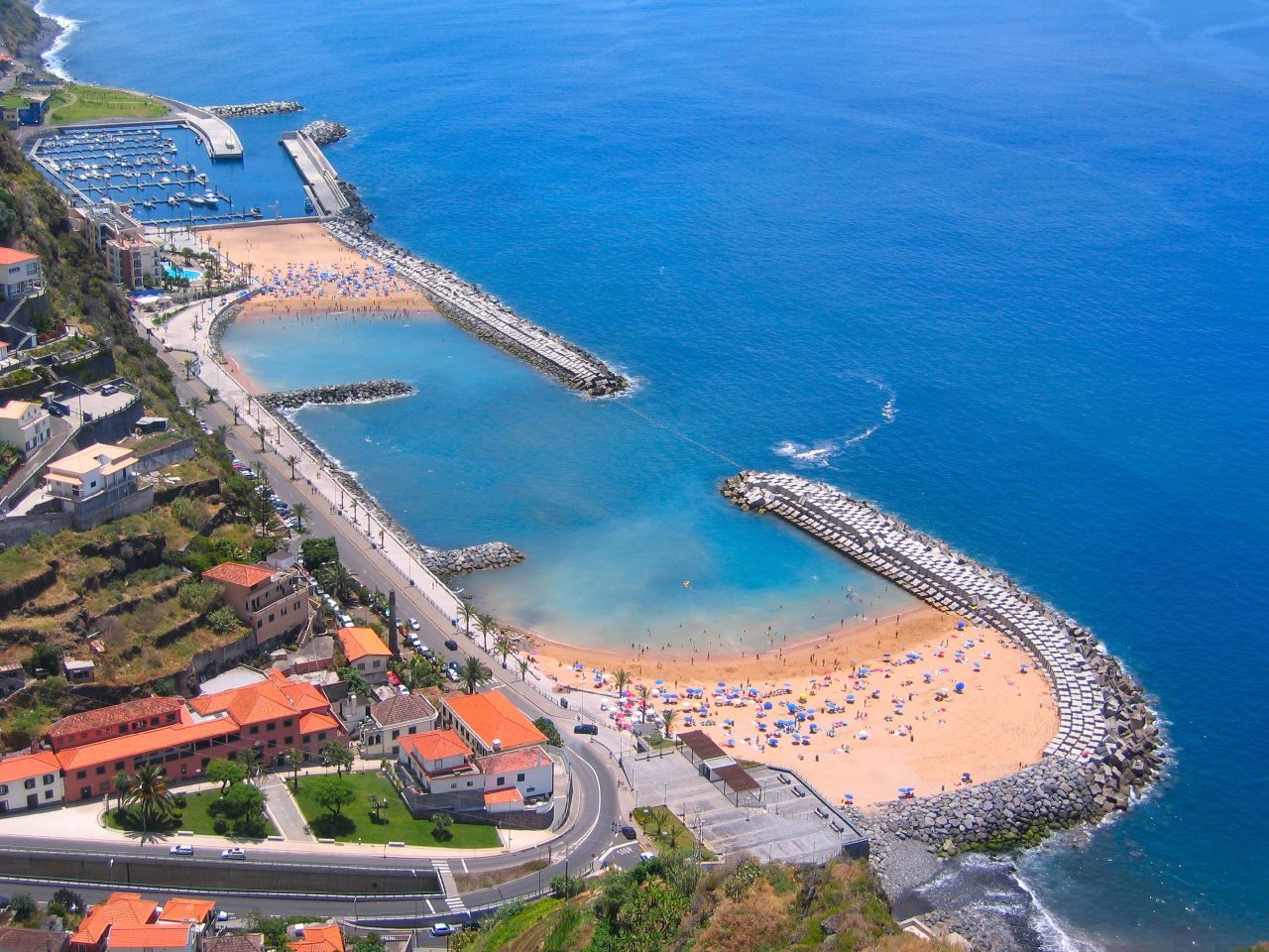 An aerial view of Calheta: two crescents of pale golden sand and calm shallow water enclosed by long rock breakwaters, a marina full of white boats above them and orange-roofed houses along the road.