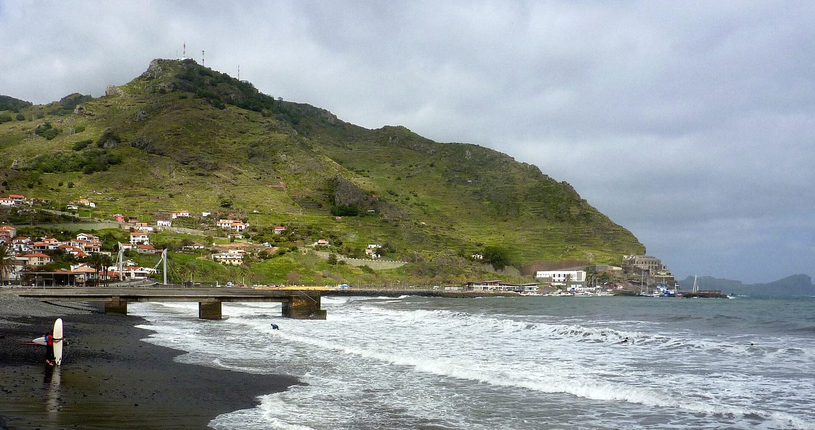 A dark shingle shore at Machico under heavy cloud, waves breaking along its length, a low concrete pier on stilts, a surfer carrying a board at the water’s edge and the green hill of Pico do Facho behind the town.