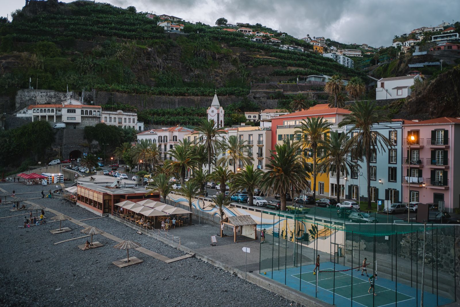 Straw parasols and loungers set out on a grey cobble beach at Ponta do Sol at dusk, restaurant terraces and a floodlit ball court along the front, pastel houses and a church tower below banana terraces.