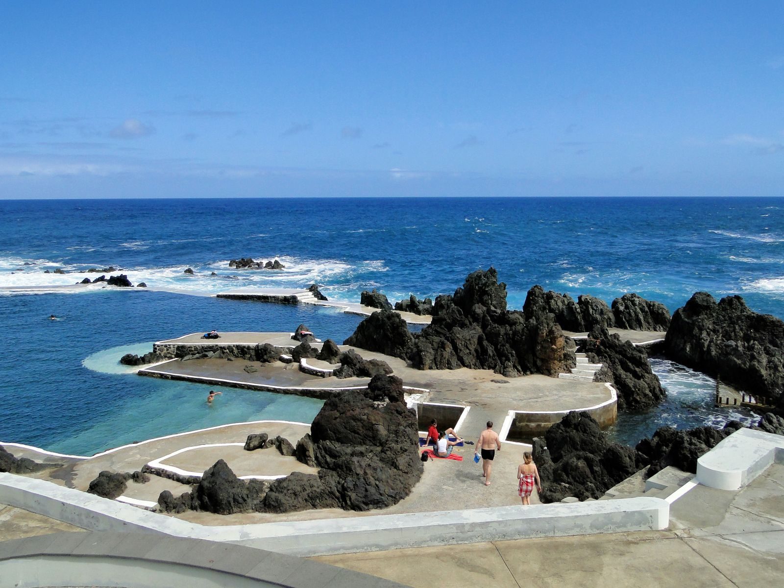 Sea-water pools among black lava outcrops behind concrete walkways at Porto Moniz, a handful of people on the paving and one swimmer in shallow turquoise water, the open Atlantic breaking beyond.
