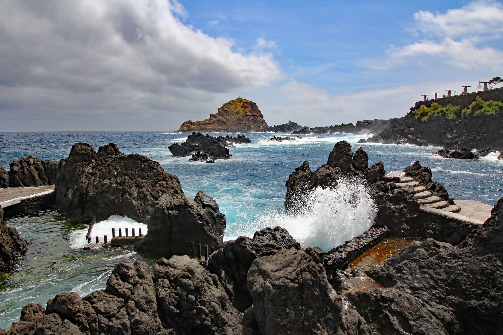 A white wave bursting over a black lava ridge into a rock pool at Porto Moniz under broken cloud, with the flat-topped Ilhéu Mole islet standing offshore.