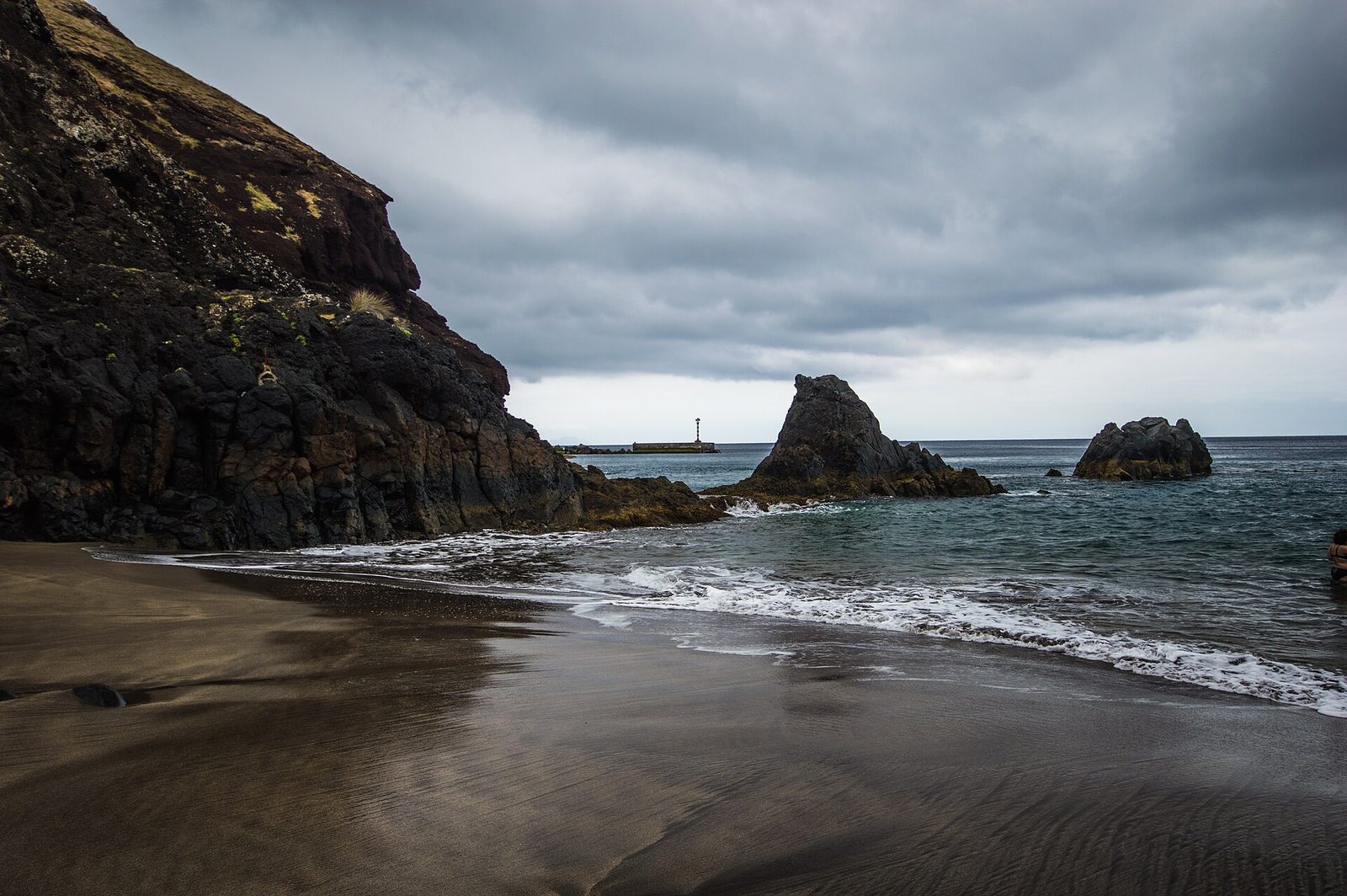 A curve of dark brown-grey sand at Prainha under storm cloud, a red-brown volcanic headland to the left, lava stacks standing in the shallows and a harbour beacon in the distance.
