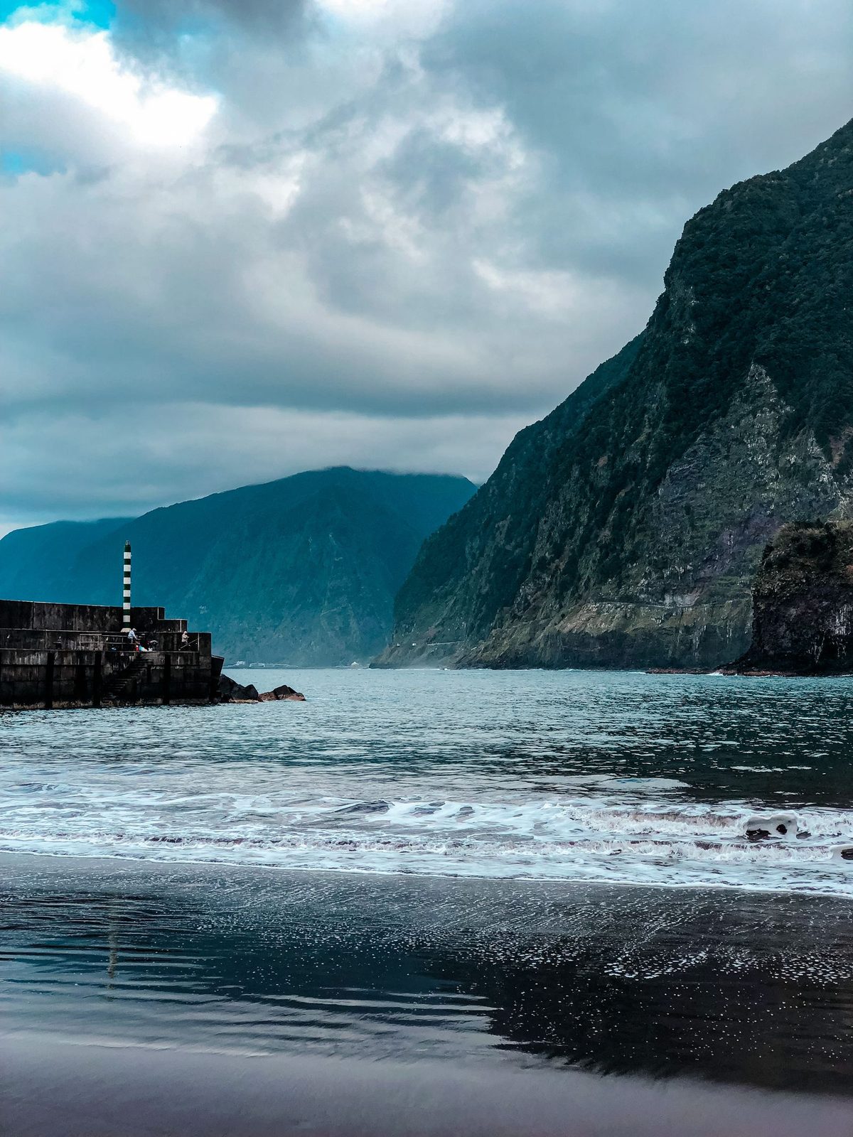 Wet black sand reflecting a grey sky at Seixal, small waves running up the beach, a stone breakwater with a striped beacon to the left and a steep green cliff dropping into the sea on the right.