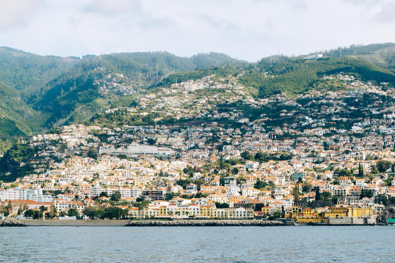 Funchal seen from the water, the city stacked up the green hillside in tiers of white walls and orange roofs, with the seafront and a fort along the shoreline.