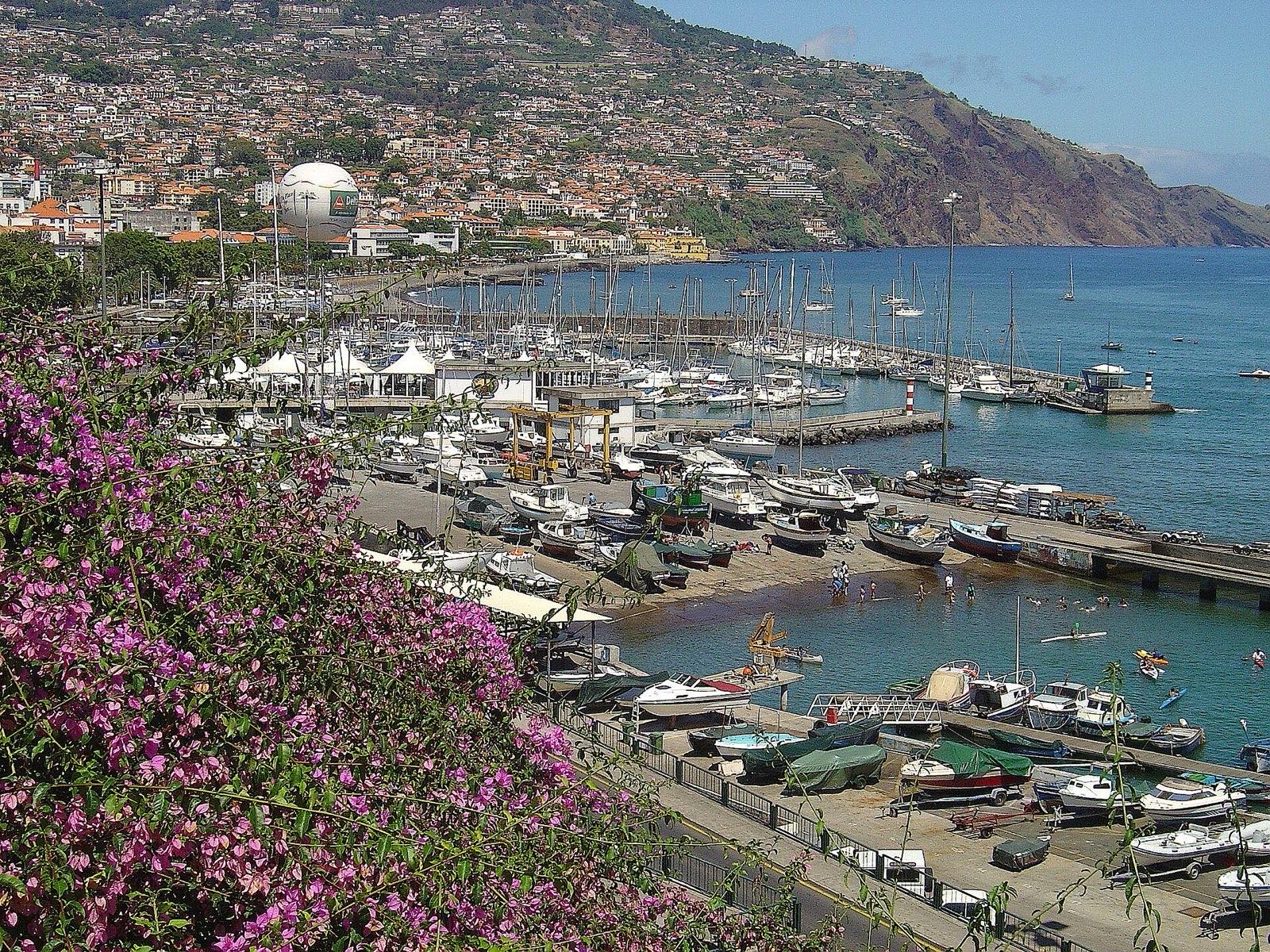 Funchal harbour from above: yachts in the marina basin, small fishing boats hauled out on the quay beside a boat lift, the city rising up the slope behind and a headland closing the far side of the bay.