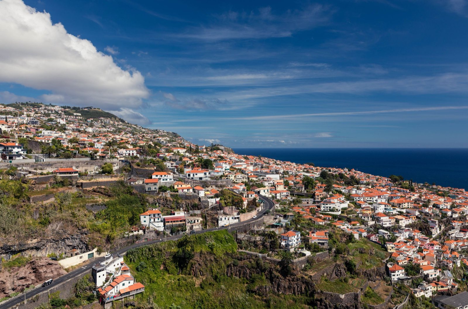 A sunlit town on the Madeiran south coast, orange roofs packed along a ridge above dark volcanic cliffs, a coast road curving through the foreground.