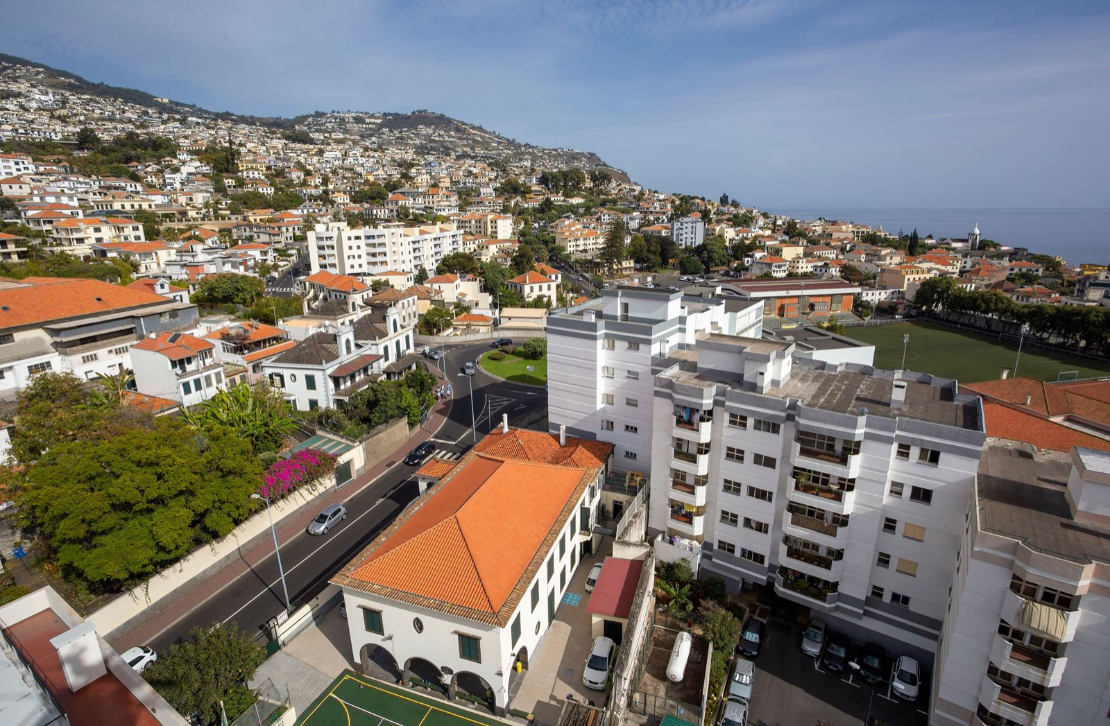 An elevated view across a Funchal neighbourhood of orange-tiled villas and white apartment blocks, the hillside suburbs climbing behind and the sea on the horizon.