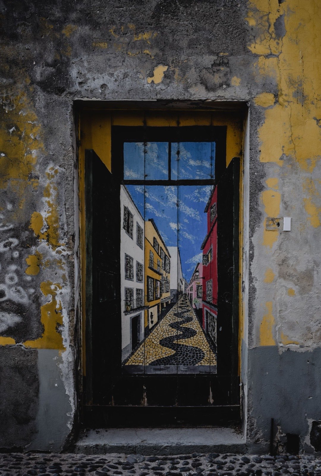 A weathered wooden door painted with a view down a Madeiran street — white, yellow and red houses above a black-and-white cobbled pavement — set in a peeling yellow wall.