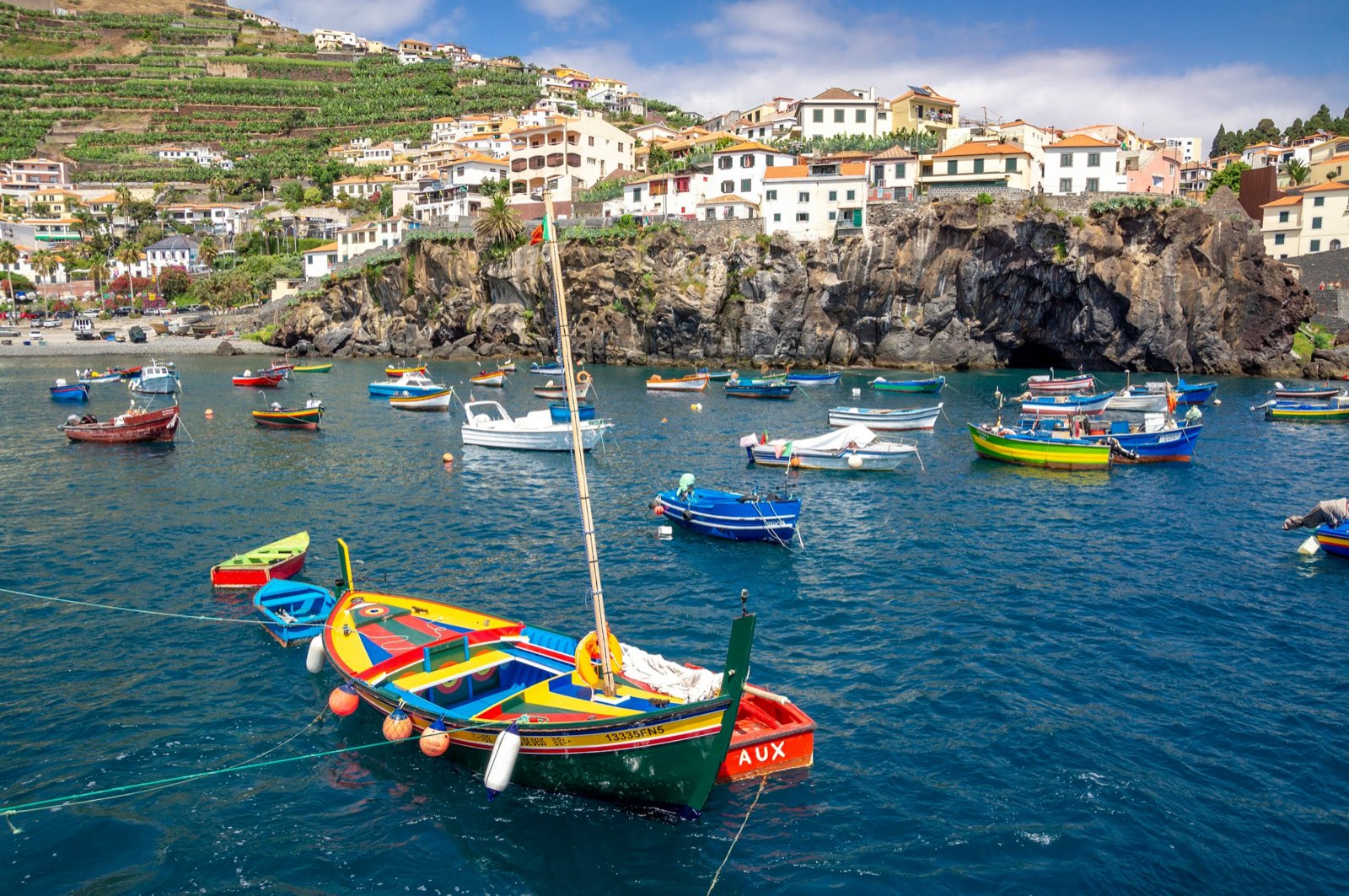 Brightly painted wooden fishing boats moored in the blue bay of Câmara de Lobos, the whitewashed town and terraced slopes rising behind a dark volcanic cliff.