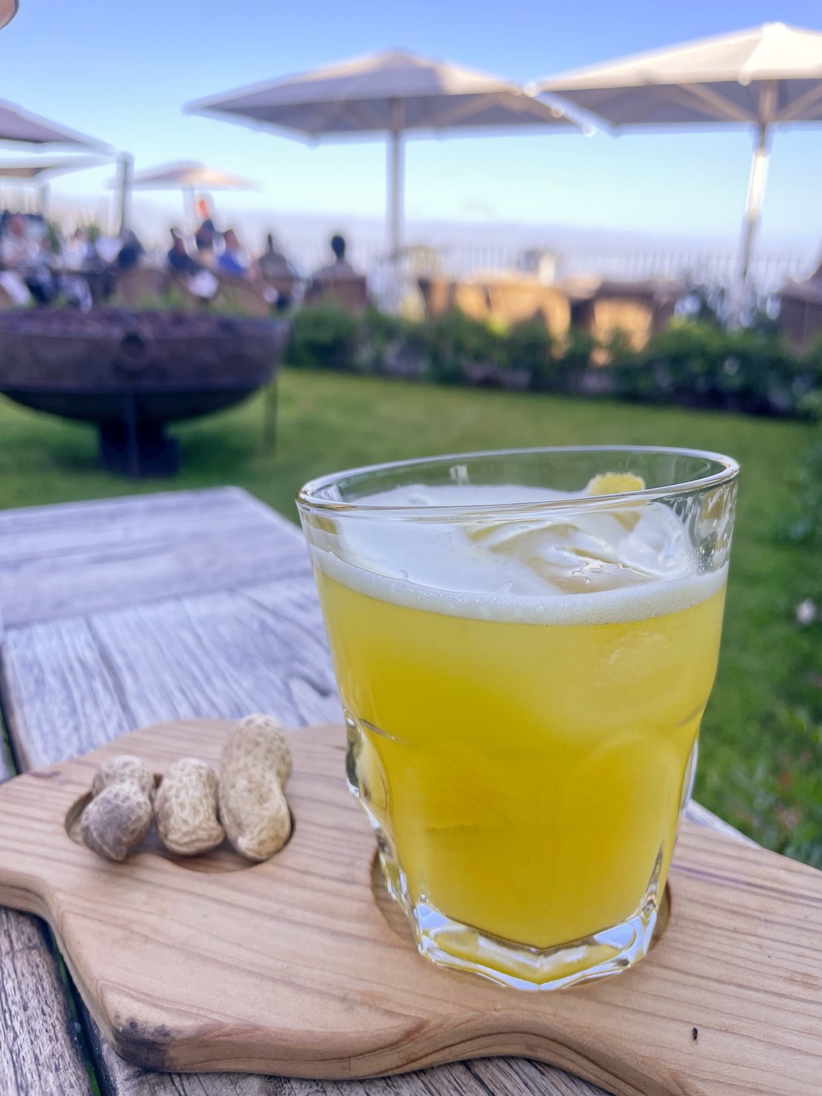 A tumbler of cloudy yellow poncha with a foamed head on a wooden board beside peanuts in their shells, on a terrace table with the sea behind.