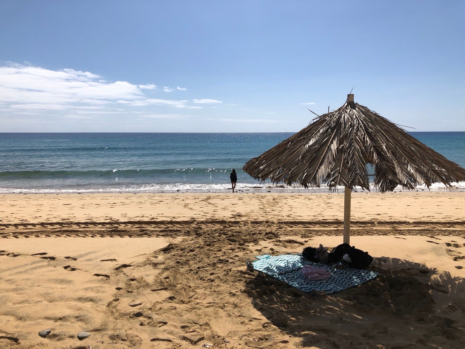 A palm-thatch parasol and a towel on wide, empty golden sand at Porto Santo, one person standing at the water’s edge and blue Atlantic beyond.