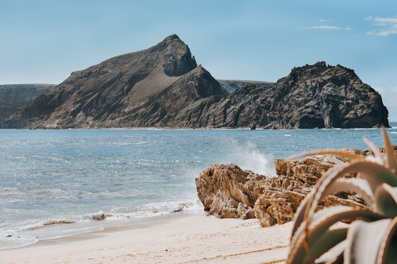 The southern end of Porto Santo’s sand, with surf running up the beach past a honey-coloured rock outcrop towards a high brown headland and an offshore stack.