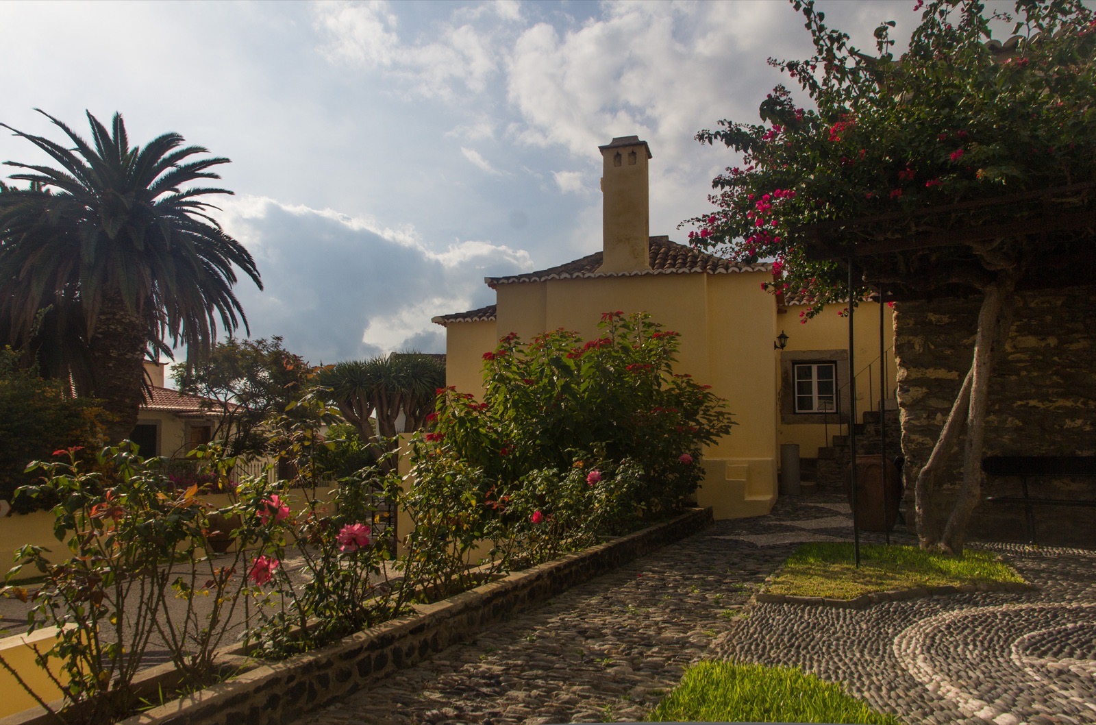 The cobbled garden path at Casa Colombo in Vila Baleira, running past roses and bougainvillea to a pale yellow house with a tiled roof and a tall white chimney.