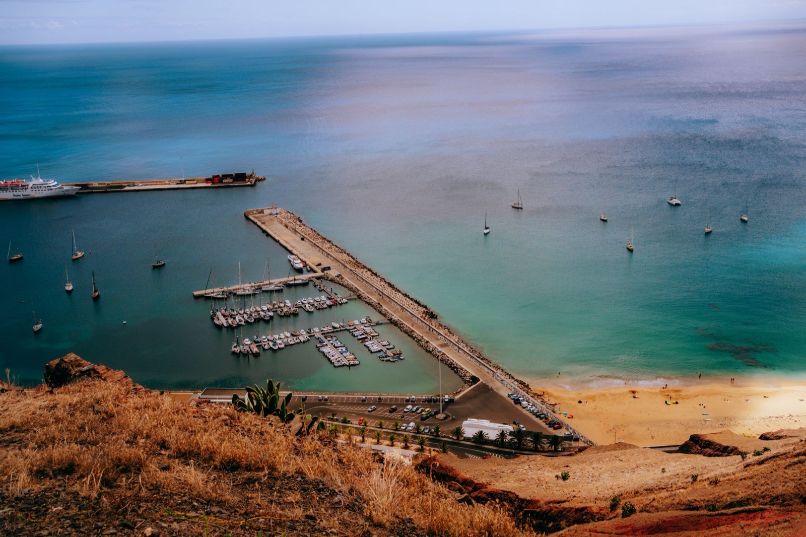 Porto Santo harbour from the hill above: a white ferry lettered Porto Santo Line at the long outer quay, a marina of yachts inside the breakwater, and the beach starting at the right.