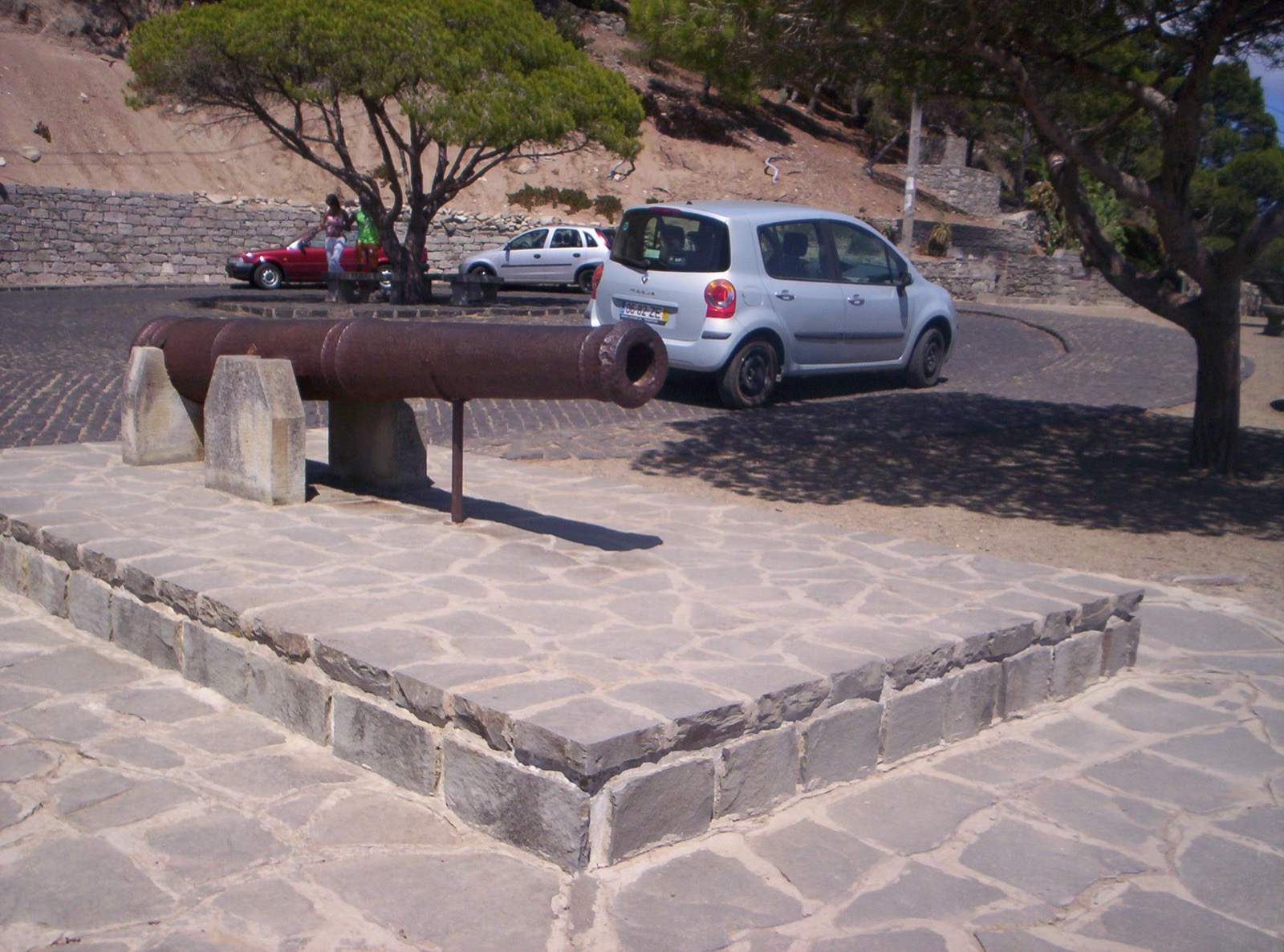 A rusted iron cannon on a stone platform at the Pico do Castelo viewpoint, with parked cars, umbrella pines and the dry hillside behind.