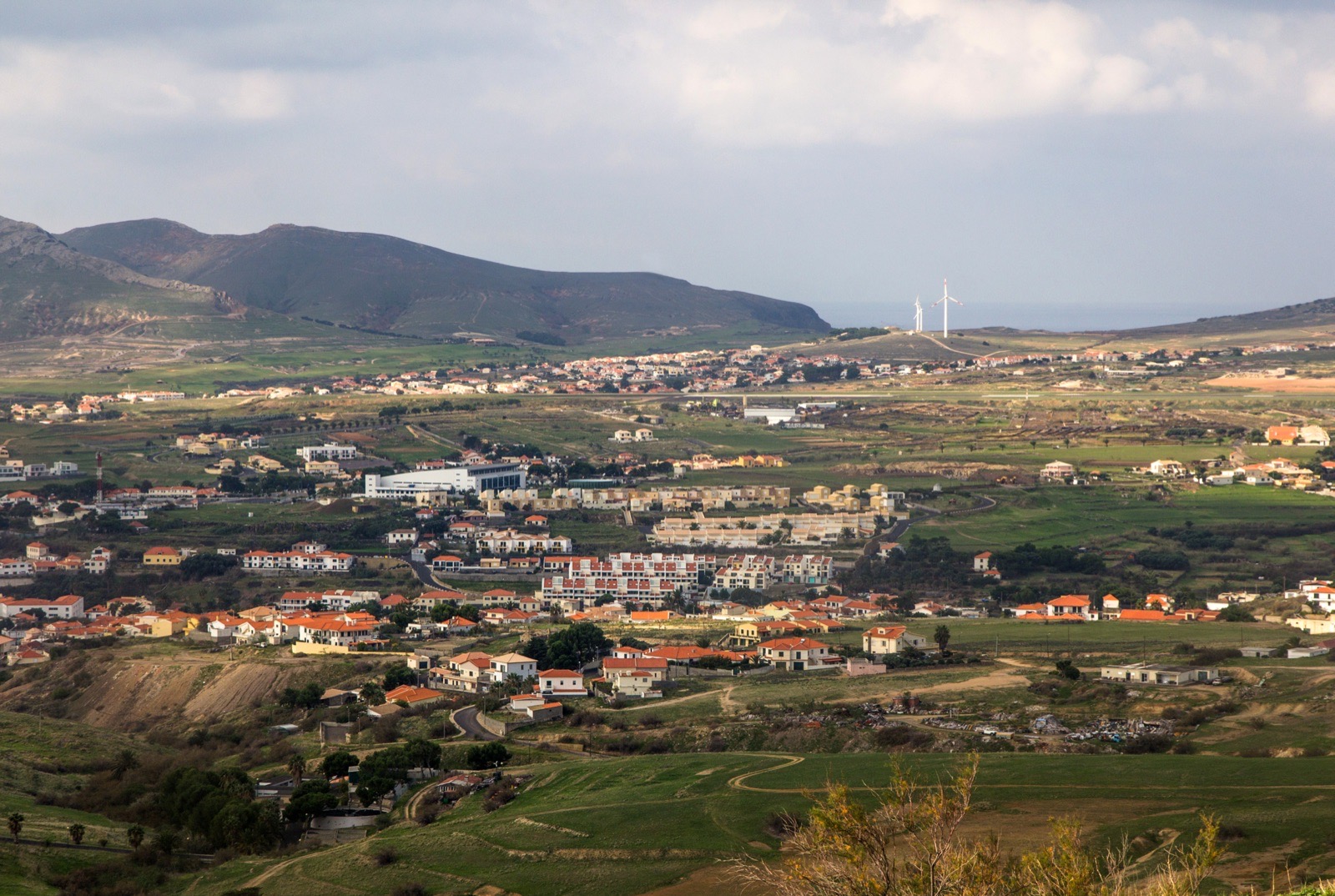 A high view across Vila Baleira: white and orange-roofed houses spread over a green plain, the airport runway and two wind turbines on the ridge behind, and bare hills above.