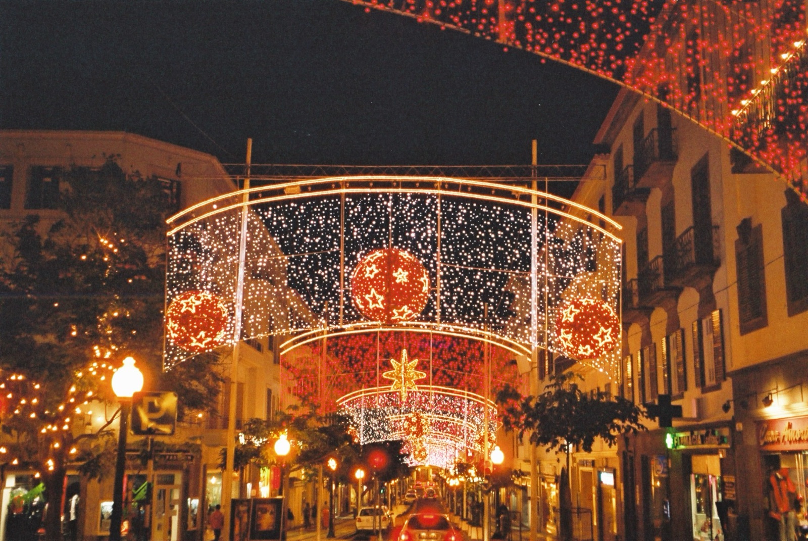 Funchal at night under strings of Christmas lights, illuminated figures above a busy street and shop windows lit behind the crowd.